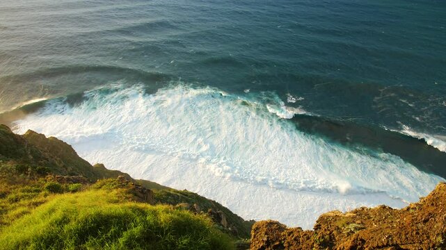 Sunny rocky coastline with green plants in mand large waves crashing against the shore turbulent ocean turbulent with white foam breaking waves