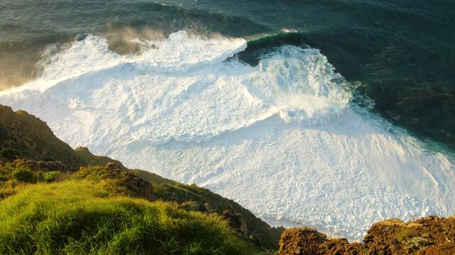 Sunny Rocky coastline with green plants in mand large waves crashing against the shore turbulent ocean turbulent with white foam breaking waves