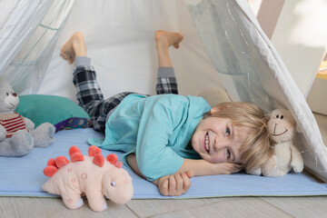 portrait of a happy 7 year old boy lying on his stomach in a wigwam with a plush dinosaur. tenderness of childhood, playing with soft toys, imaginary friend. Cozy interior of a children's room © Anna