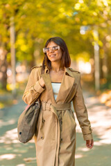 A woman wearing a tan trench coat and sunglasses walks down a sidewalk