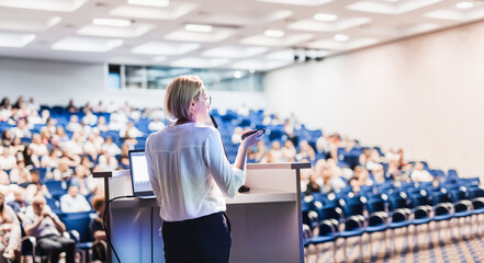 Female speaker giving a talk on corporate business conference. Unrecognizable people in audience at...