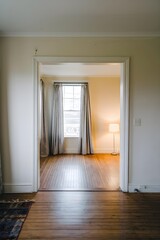 an empty room with a hardwood floor and white walls. There is a window with curtains on the left side, and a doorway with a light switch on the right side. The room has a lamp.