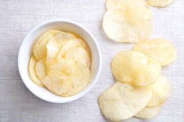 Salted potato chips in a white bowl on linen fabric. Fresh and ready to eat crisps, slim slices of deep fried potato, made from tubers of the root vegetable Solanum tuberosum. Close-up, from above.