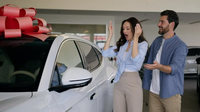 Caucasian lovely man male guy husband boyfriend make surprise present gift close eyes to woman female girl wife girlfriend amazed wonder happy couple buying new car hugging auto in showroom automobile
