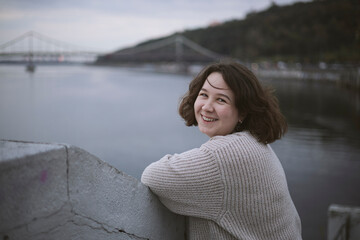 Fototapeta premium Portrait of happy young woman near river on city bridge in cold cloudy weather.