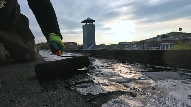 a man priming a roof before repairing the roofing felt, to avoid leaks in the future, against the backdrop of a sunset in an urban development