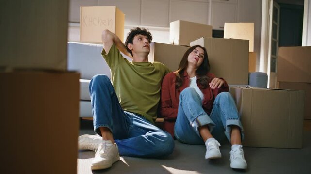 Tired couple moving house sitting floor modern living room between carton boxes
