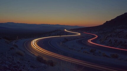 As the sun sets on the horizon a line of cars can be seen following the winding curves of the desert highway their lights creating a beautiful contrast against the dusky sky.