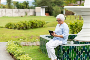 Fototapeta premium Asian senior women using computer tablets technology for entertainment, living in a retirement home facility outside park nature, Happy smiling joyful.