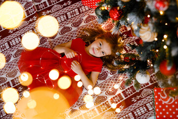 A joyful girl in a red dress is sprawled beneath the Christmas tree, grasping a garland, viewed...
