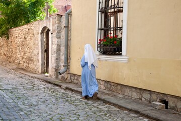 The photo shows a nun in white and blue clothes walking along an old building. Her calm and smooth gait creates an atmosphere of peace