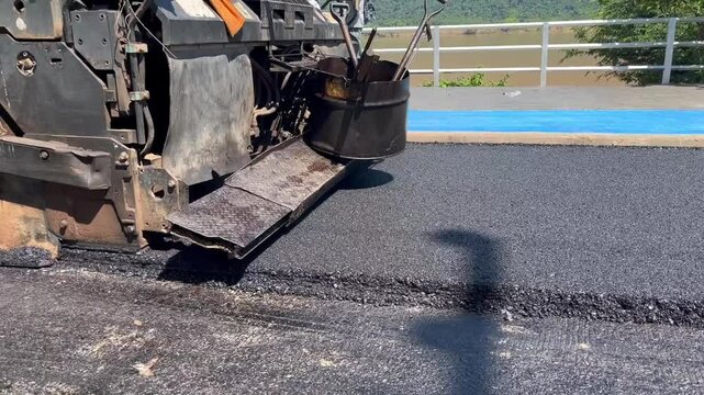 asphalt paver machine laying fresh asphalt on a road. The image captures the machinery in action, showing the spreader screed and other components, smooth layer of asphalt, construction site, civil 