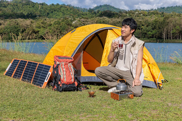 An Asian man sitting and drinking coffee near camping tent beside the lake and mountain in the sunshine morning with backpack and mobile solar cell
