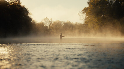 Young fly fisherman casting on a foggy river at sunrise