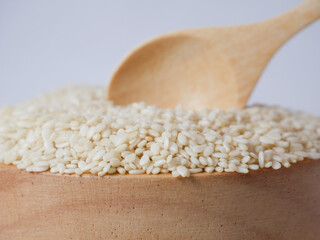 Close-up photograph of white sesame seeds in a wooden bowl accompanied by a small wooden spoon, placed on a clean white background. Perfect for illustrating healthy food ingredients, cooking concepts