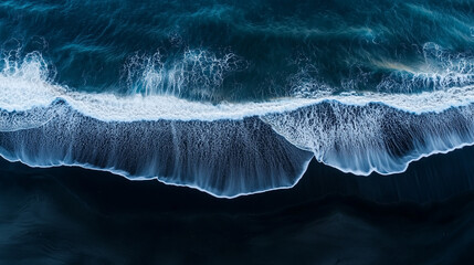 Aerial view of the sea and beach, with waves crashing onto black sand. The ocean is deep blue in color, creating an endless horizon. 