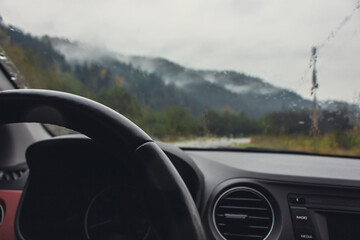 Driving in mountains on rainy day. Car dashboard with steering wheel with rain drops on glass and...