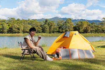 An Asian man sitting and looking at the camera while drinking coffee near camping tent beside the lake and mountain in the sunshine morning with backpack and mobile solar cell
