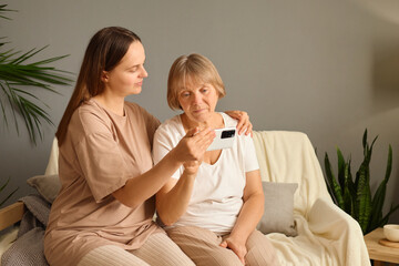 Senior woman sitting on couch with adult daughter teaching her about technology scrolling on...
