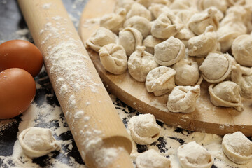 Raw dumplings, selective focus. Cooking pelmeni, flour, eggs and wooden rolling pin. Meat pelmeni or ravioli on wooden board, dark table. Slavic traditional dish. Food background