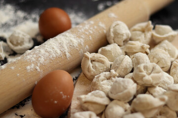 Raw dumplings, selective focus. Cooking pelmeni, flour, eggs and wooden rolling pin. Meat pelmeni or ravioli on wooden board, dark table. Slavic traditional dish. Food background