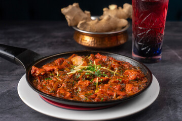 Sizzling dish of mixed beef meats, spiced sauce, garnished with fresh herbs. In the background, a textured glass filled with a deep red drink, alongside a copper bowl of fresh ginger.