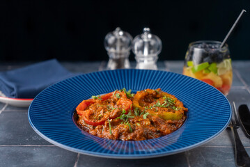 Spicy Indian red beef curry, garnished with bell peppers, and fresh herbs, served on a blue textured plate. a colorful glass of fruity drink, the table is set with a folded navy napkin.