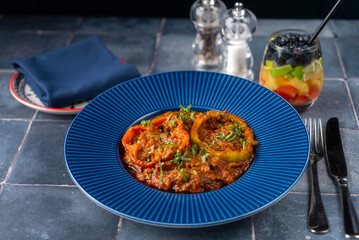 Spicy Indian red beef curry, garnished with bell peppers, and fresh herbs, served on a blue textured plate. a colorful glass of fruity drink, the table is set with a folded navy napkin.