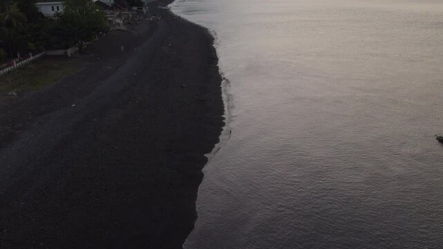 Atardecer en una playa de arena negra y palmeras en Bali d&oacute;nde el sol se pone detr&aacute;s de un volc&aacute;n y nubes bajas. Barco atracado en la orilla y personas en la arena.