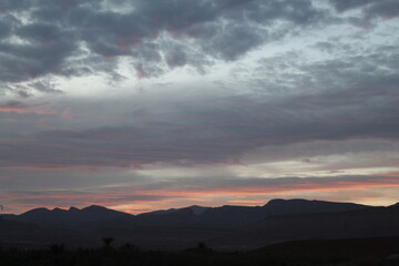 A panoramic view of a mountainous landscape during dusk. The sky is filled with dramatic clouds, and faint pink and orange hues near the horizon cast a serene ambiance over the distant hills
