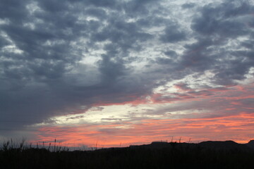 A landscape view of a sunset with vibrant red and orange hues in the sky,  covered by dark, fluffy clouds. The silhouette of distant hills and grassy foreground adds depth to this creative work