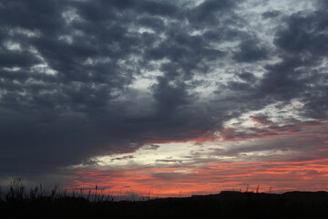 A dramatic sunset with dark, heavy clouds covering the sky becomes a creative work of art. The horizon glows with shades of red, orange, and pink, a striking contrast against the silhouetted landscape
