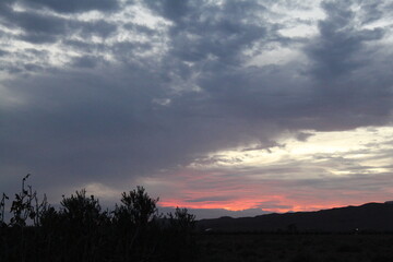 A dramatic sky with dark clouds partially obscures the setting sun, while hints of pink and orange appear on the horizon, silhouetting distant hills and foliage.