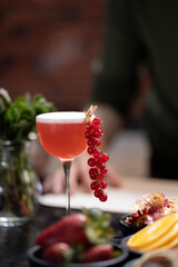 Red cocktail garnished with fresh red currants in tall glass, placed on rustic table with various fruits and greenery in the background, evoking refreshing, vibrant, and summery atmosphere, copy space