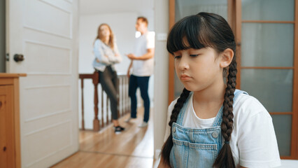 Stressed and unhappy young girl huddle in corner, cover her ears blocking sound of her parent arguing in background. Domestic violence at home and traumatic childhood develop to depression. Synchronos
