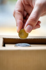 Close-up of a hand placing a coin in a cardboard donation box. Donations, volunteering, foundation. Simple background with copy space