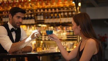 Sitting in smiling beautiful woman guest lively taking with smart bartender while drinking mixed vodka in blue hawaii cocktail and whiskey at luxurious counter bar on Friday meeting at club. Vinosity.