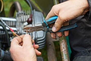 worker holds old iron mechanical industrial sharp blue plastic handle dirty nippers in hand and cuts protective copper electrical wire during repair work outdoors during daytime