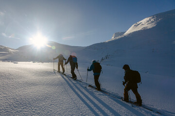 A group of climbers ascends a steep, snowy slope of big rocky moutn under a clear, blue sky.