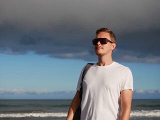 A young man with a backpack, wearing a white T-shirt and sunglasses standing on the seashore at the background of gloomy cloudy sky, with copy space. Tourism, traveling and resort recreation concept