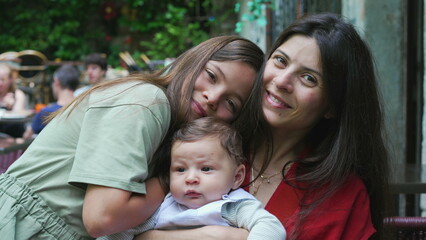 Naklejka premium Mother and her two children sharing a warm embrace outdoors. Young girl hugs her mother and baby sibling, expressing love and closeness in a relaxed and joyful family moment