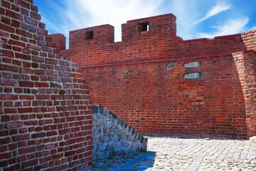 A historic red brick fortress wall with two turrets stands under a clear blue sky. A charming cobblestone street leads into the distance, featuring a hole in the larger turret. © Ilja