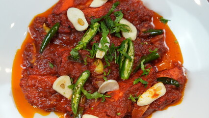 Flavorful dish marinated meat, glistening in a rich red sauce made with spices and tomatoes. garnish with fresh green chilies, garlic slices, and a cilantro sprinkle.