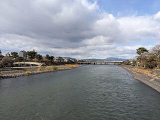 Uji, Japan - 12.17.2023: View of Uji Bridge across Uji River alongside riverside footpaths and trees in an autumn afternoon under a blue sky with clouds