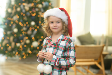 Kid boy in santa hat near christmas tree at home.