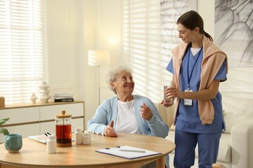 Fototapeta premium Caregiver giving glass of water to senior woman indoors. Home health care service