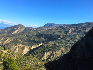 Obraz premium Aerial view of the foothills from the top of the mountain. Old olive groves, green fields, road bends among the slopes, mountain peaks under the blue sky. Greece, Peloponnese.