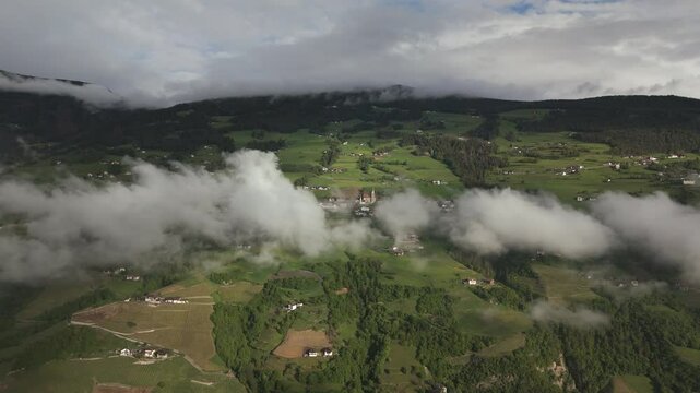 Aerial footage of a mountain valley with patches of clouds drifting over green fields and small villages, surrounded by forested hills.