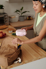Young Hispanic woman preparing lunch in modern kitchen, placing meal container in her bag. Fresh...