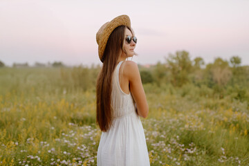 a woman in a long light dress stands in a flowers field against the sky, her back to the camera. summer look, freedom field happiness portrait young woman